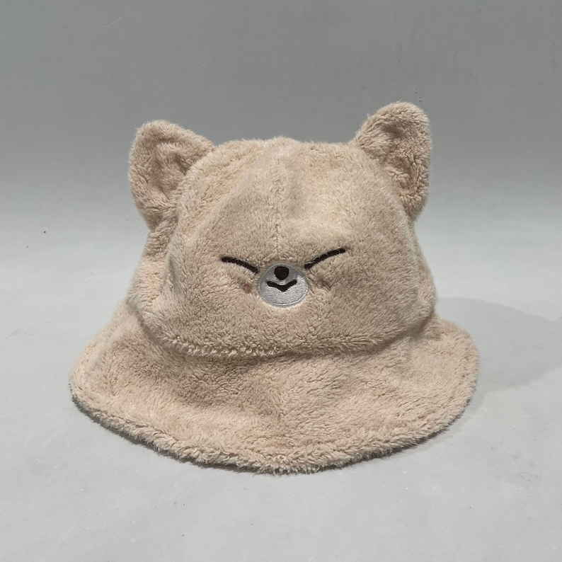 Op de afbeelding: Een beige, pluche bucket hat met een schattig geborduurd beren gezicht.