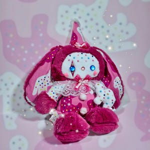 May include: A plush rabbit toy in shades of magenta and pink, featuring a cone-shaped party hat, a white lace collar, and blue circular eyes. The rabbit has long ears with a colorful confetti pattern and a small white plush figure attached to its side.