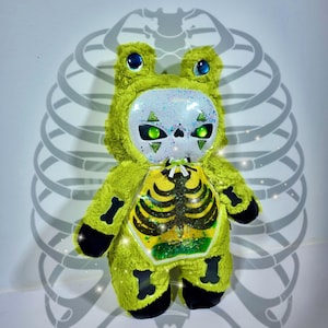 May include: A plush frog toy with lime green fur, featuring a white glittery face mask with green eyes and triangular markings. The chest area has a clear window displaying a black skeleton ribcage graphic, and black bone patches on the limbs.