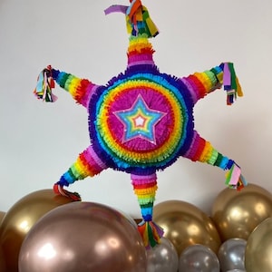 May include: A rainbow-coloured star-shaped pinata with a pink star in the centre, surrounded by gold, silver and white balloons.