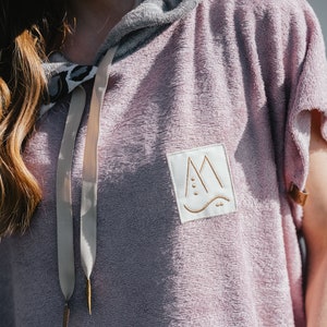 May include: A hooded towel in a light purple color with a white patch on the front. The patch has a gold outline and a stylized mountain design with a wave at the bottom. The towel has a light gray hood and a beige drawstring.