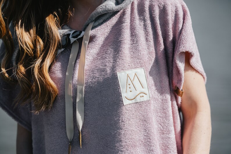 May include: A hooded towel in a light purple color with a white patch on the front. The patch has a gold outline and a stylized mountain design with a wave at the bottom. The towel has a light gray hood and a beige drawstring.