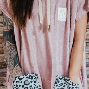 May include: A pink hooded towel with a leopard print lining the pockets. The towel has a small logo with the letter "M" and a wavy line.