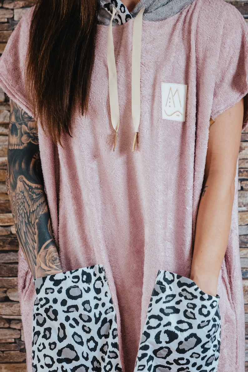 May include: A pink hooded towel with a leopard print lining the pockets. The towel has a small logo with the letter "M" and a wavy line.