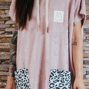 May include: A pink hooded towel with a gray hood and a small patch with a mountain design. The towel has two large pockets with a leopard print lining.