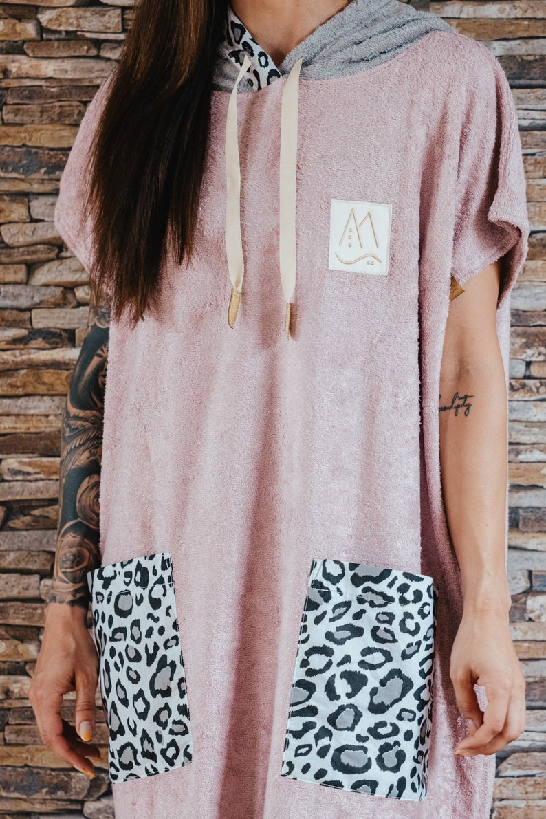 May include: A pink hooded towel with a gray hood and a small patch with a mountain design. The towel has two large pockets with a leopard print lining.