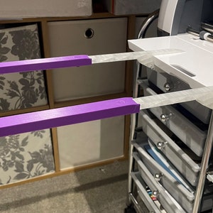 May include: Two purple 3D printed arms attached to a white machine with a silver handle. The machine is on a metal trolley with drawers.