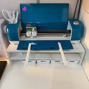 May include: A teal blue electronic cutting machine with a white stand and four white storage bins. The machine has a lid that is open and a cutting blade is visible.