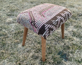 May include: A brown and pink patterned footstool with wooden legs. The footstool is sitting on a patch of grass.