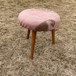May include: A pink and brown patterned stool with wooden legs. The stool is sitting on a patch of brown grass.