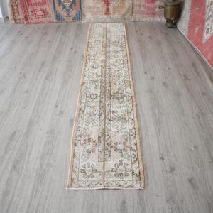 Pode incluir: Um tapete corredor longo e estreito de cor bege com um padrão floral tradicional. O tapete tem um aspeto desbotado e vintage e é feito de um material macio e tecido.