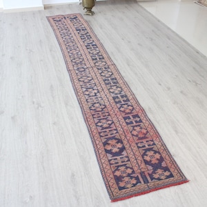 May include: A long, rectangular runner rug with a repeating geometric pattern in navy blue, red, and beige. The rug is placed on a light-colored wooden floor. The rug's design features stylized floral and geometric shapes.