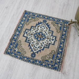Op de afbeelding: Een klein, vierkant, handgeknoopt tapijt met een centraal geometrisch ruitvormig medaillon in tinten blauw, beige en wit. Het tapijt heeft een dikke blauwe rand met ingewikkelde patronen en korte, lichtgekleurde franjes langs de randen.