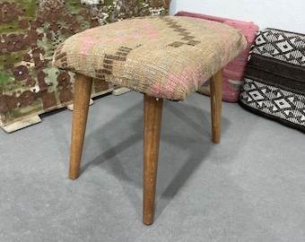 May include: A small, wooden stool with a woven fabric seat in shades of brown, pink, and white. The stool has four legs and is sitting on a gray surface.