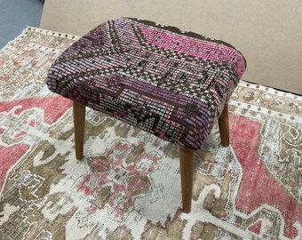 May include: A small, square ottoman with a brown, pink, and purple patterned fabric top and wooden legs. The ottoman is sitting on a rug with a similar pattern.