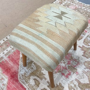 May include: A light brown and white patterned footstool with wooden legs. The footstool is sitting on a rug with a pink and white pattern.