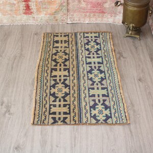 May include: Rectangular rug with a central design of vertical columns in navy blue and tan. The rug has a fringed edge and is placed on a light wood floor. The rug's design includes geometric shapes and floral motifs.