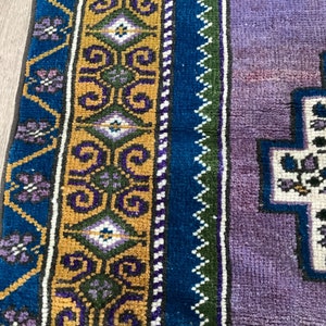May include: A close-up of a colorful rug with a geometric pattern. The rug has a purple, blue, green, and gold border. The center of the rug is white with a floral design.
