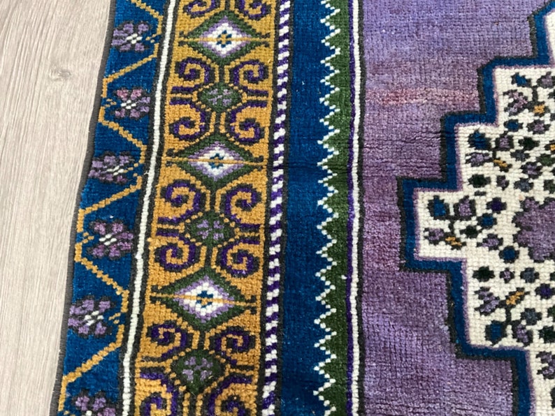 May include: A close-up of a colorful rug with a geometric pattern. The rug has a purple, blue, green, and gold border. The center of the rug is white with a floral design.