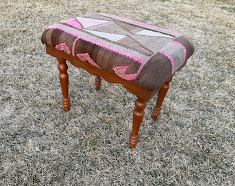 May include: A small, rectangular wooden stool with a patterned fabric seat. The seat features a patchwork design in shades of brown, pink, and gray. The legs are turned and stained a warm brown color, adding a touch of elegance to the piece.