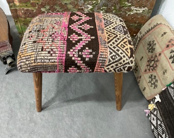 May include: A small, wooden stool with a colorful, patterned fabric seat. The fabric is a mix of pink, brown, and white, and features geometric designs. The stool has four legs and is made of dark wood.
