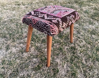 May include: A small, brown and pink patterned ottoman with wooden legs. The ottoman is sitting on a patch of brown grass.