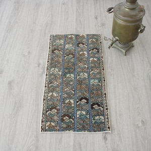 May include: A rectangular rug with a detailed pattern in shades of blue, brown, and beige. The rug is placed on a light-colored wooden floor. A vintage brass samovar is in the background.