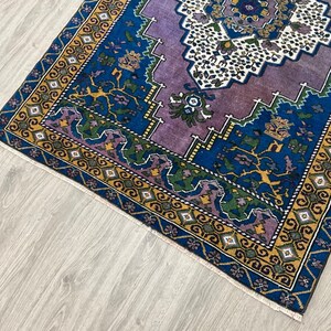 May include: A vintage rug with a purple, blue, and yellow geometric pattern. The rug has a fringed edge and is made of wool.