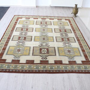 May include: A rectangular area rug with a cream-colored field and geometric patterns in shades of brown, gold, and gray. The rug has a border with a repeating design and is placed on a light-colored wooden floor.