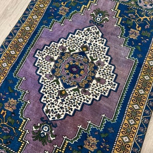 May include: A vintage rug with a purple center and blue and yellow borders. The center features a geometric design with white and green accents.