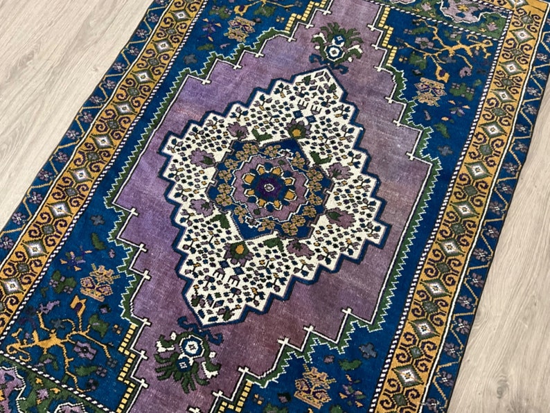 May include: A vintage rug with a purple center and blue and yellow borders. The center features a geometric design with white and green accents.