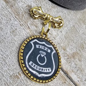 以下が含まれることがあります： 黒と白のエナメルチャームが付いたゴールドトーンのブローチ。チャームは盾の形をしており、「リングセキュリティ」という言葉とダイヤモンドリングのグラフィックが描かれています。