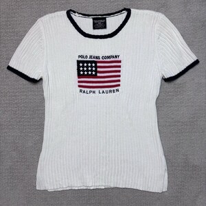 Peut inclure: Un t-shirt blanc à manches courtes et texture côtelée, doté d'un liseré bleu foncé au col et aux manches. Le centre présente un drapeau américain brodé avec le texte POLO JEANS COMPANY au-dessus et RALPH LAUREN en dessous.