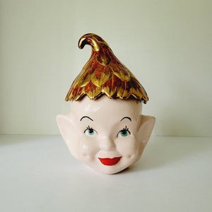 May include: A ceramic elf head cookie jar with a gold and red leaf design on the hat. The elf has a pink face, blue eyes, and a red smile.