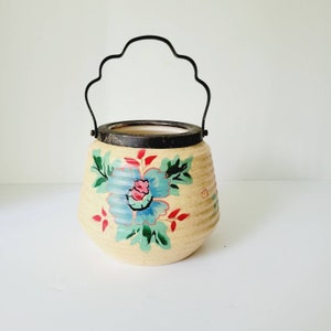 Op de afbeelding: Een vintage keramische koekjespot met een bloemenmotief. De pot heeft een crèmekleurige basis met een blauw, roze en groen bloemenpatroon. Hij heeft een zwart metalen handvat en een zilverkleurig deksel.