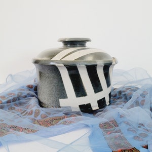 May include: A ceramic storage jar with a black and white geometric pattern. The jar is sitting on a blue and white patterned fabric.