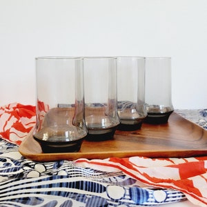 May include: Four clear glass tumblers with a dark gray base, sitting on a wooden tray. The tray is on top of a colorful patterned fabric.