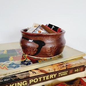 Puede incluir: Un cuenco de cerámica marrón hecho a mano, con una superficie texturizada y diseños negros y de sol. El cuenco está lleno de pequeños objetos coloridos y descansa sobre una pila de libros con títulos como "Art in America" y "Making Pottery".
