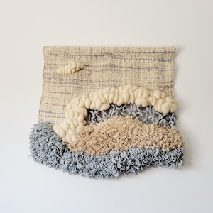 May include: A beige and gray woven wall hanging with a textured, fluffy design. The hanging features a cloud-like shape with a raised, fluffy texture.