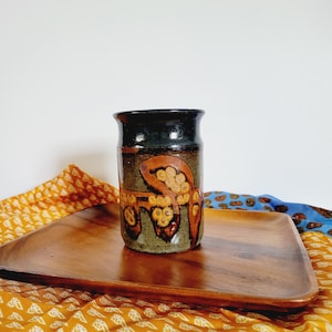 Puede incluir: Un jarrón de cerámica marrón con un esmalte azul oscuro y un diseño pintado a mano de hojas naranjas y amarillas. El jarrón está sobre una bandeja de madera.