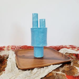 Puede incluir: Un jarrón de cerámica azul claro con un diseño apilado único. El jarrón tiene dos picos cilíndricos y una sección central más ancha, colocado sobre una bandeja de madera. La bandeja está colocada sobre una tela estampada con diseños florales rojos y marrones.