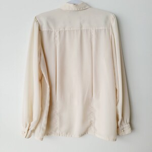 Op de afbeelding: Cr&egrave;mekleurige blouse met lange mouwen, een knoopsluiting en een klassieke kraag. De blouse hangt aan een witte hanger tegen een witte achtergrond. De mouwen hebben manchetten met knopen.