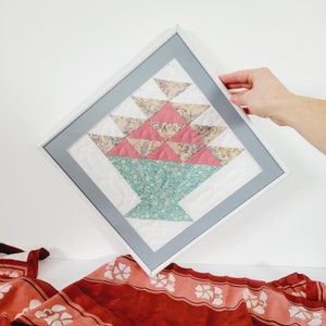 May include: Framed quilt art with a basket design. The basket is made from floral patterned fabrics in pink, beige, and teal. The quilt is framed in a white wooden frame with a grey mount. A unique piece of home decor.