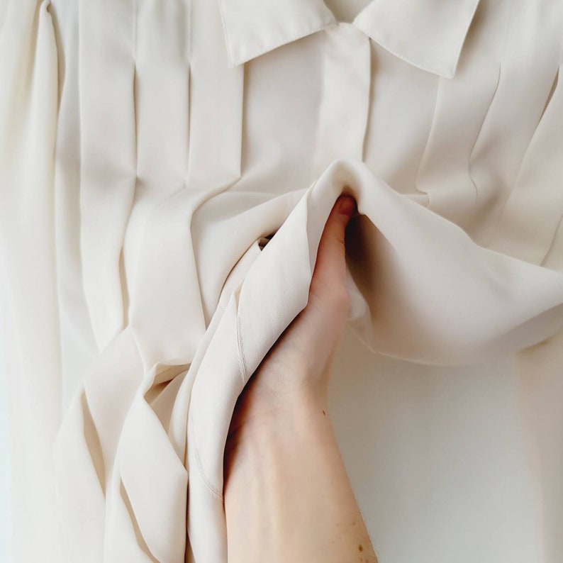 Op de afbeelding: Een cr&egrave;mekleurige blouse met een klassieke kraag en geplooide voorkant. De stof lijkt zacht en licht van gewicht, met een subtiele glans. De blouse wordt omhoog gehouden, wat de drape van het materiaal laat zien.