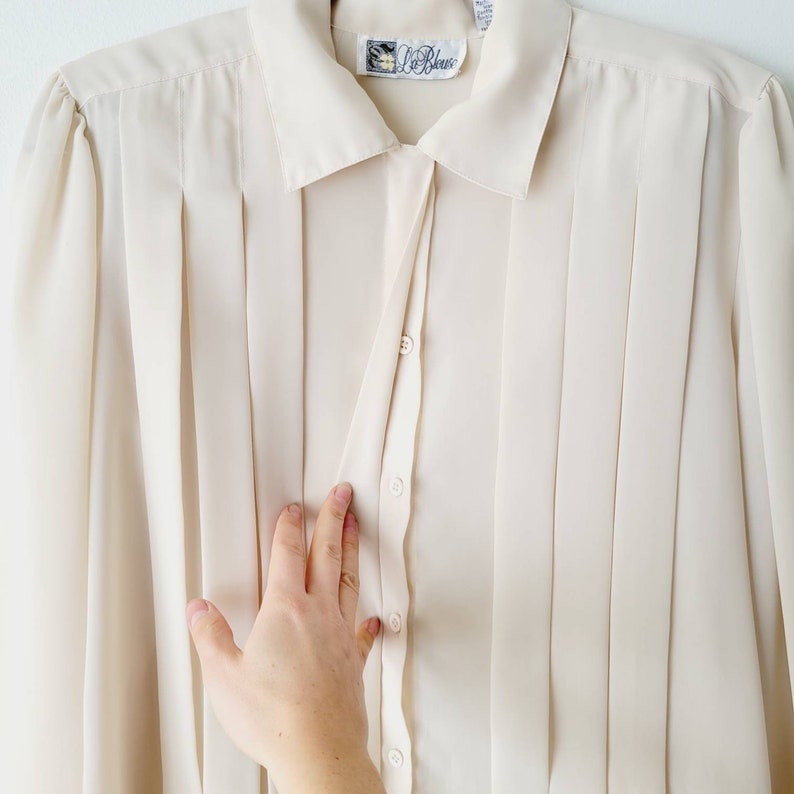 Op de afbeelding: Een cr&egrave;mekleurige blouse met knopen, verticale plooien en een kraag. Het shirt met lange mouwen heeft een label met de tekst "Le Bleu". De blouse is gemaakt van een lichte stof.