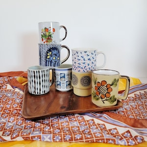 May include: A collection of six ceramic mugs with various designs, including floral, geometric, and abstract patterns. The mugs are stacked on a wooden tray, showcasing a range of colors and styles, perfect for coffee or tea.