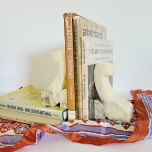 May include: A pair of off-white marble bookends supporting a stack of books. The bookends have a curved, abstract design. The books include titles like "Creative Casting" and "Abstract". A patterned scarf with red, blue, and white accents is draped in the foreground.