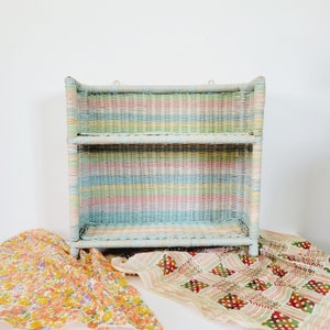 May include: A pastel-colored wicker shelf with two tiers. The shelf is painted in horizontal stripes of blue, pink, yellow, and green. The shelf has a rectangular shape and is designed to be hung on a wall. Two decorative fabrics are draped in front of the shelf.