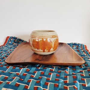 May include: A round, beige ceramic pot with orange abstract designs sits on a square wooden tray. The tray rests on a blue and red patterned fabric. The pot is a decorative item.