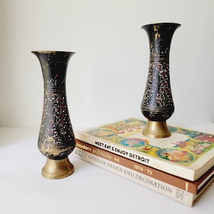May include: Two black and gold vases with intricate floral designs and red accents. The vases are sitting on top of a stack of books with the text "MEET, EAT & ENJOY DETROIT" and "modern publicity 44 1974/75 INTERIOR DESIGN AND DECORATION".